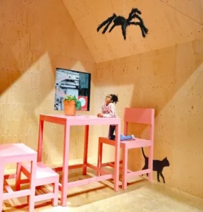 Ein Mädchen sitzt auf einem großen rosa Stuhl und schaut eine riesige Spinne an der Decke an.