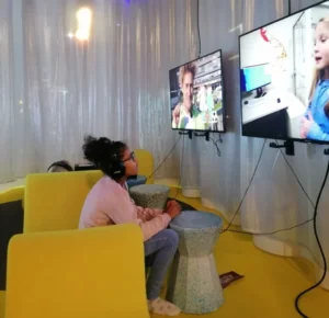 Ein Mädchen mit Kopfhörern sitzt in einem gelben Sessel vor zwei großen Bildschirmen.