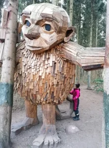 Ein Mädchen in einer rosa Jacke neben einer riesigen Holzskulptur eines Trolls im Wald.